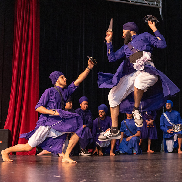 Sikh men in purple outfits performing martial arts on stage.