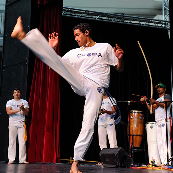 Capoeira martial artist in an all-white outfit performing a kick on stage.