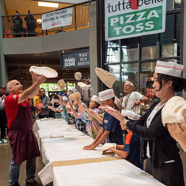 A man in front of a table of children all tossing pizza dough.