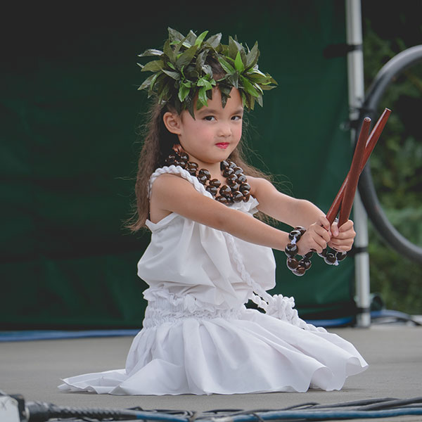Little girl in a traditional Hawaiian outfit posing with wooden sticks on stage.