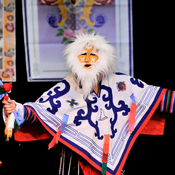 Performer wearing a colorful outfit with complex patterns, wearing a furry mask and holding a colorful, cloth-woven staff.