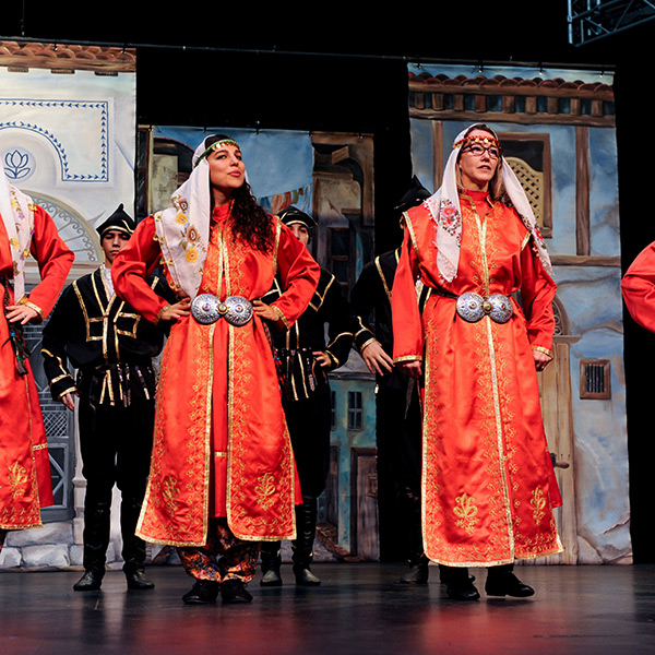 Four females performers wearing red and gold robes with white headdresses standing with hands on their hips.